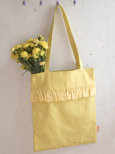 A yellow ruffle tote bag holding yellow flowers is hung on a peg behind a terrazzo wall.