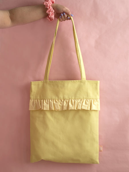A female arm holds a yellow ruffle tote bag against a pink foreground.