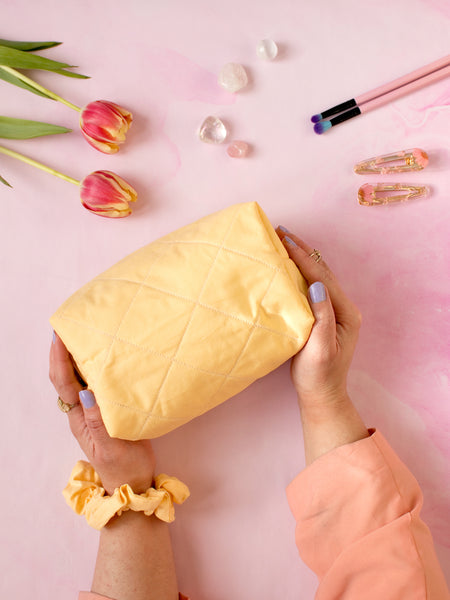 A pair of female hands holding a yellow quilted zip pouch on a pink-marbled floor. Flowers, crystals, makeup brushes and hair clips offset at the top.