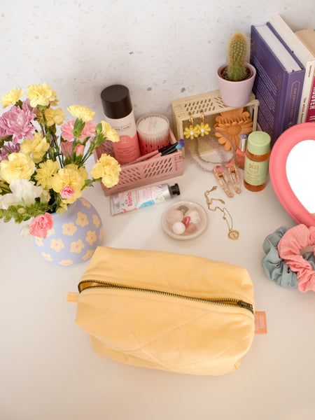 A yellow quilted zip pouch opened on a busy dressing table filled with beauty products, books and a vase of flowers.