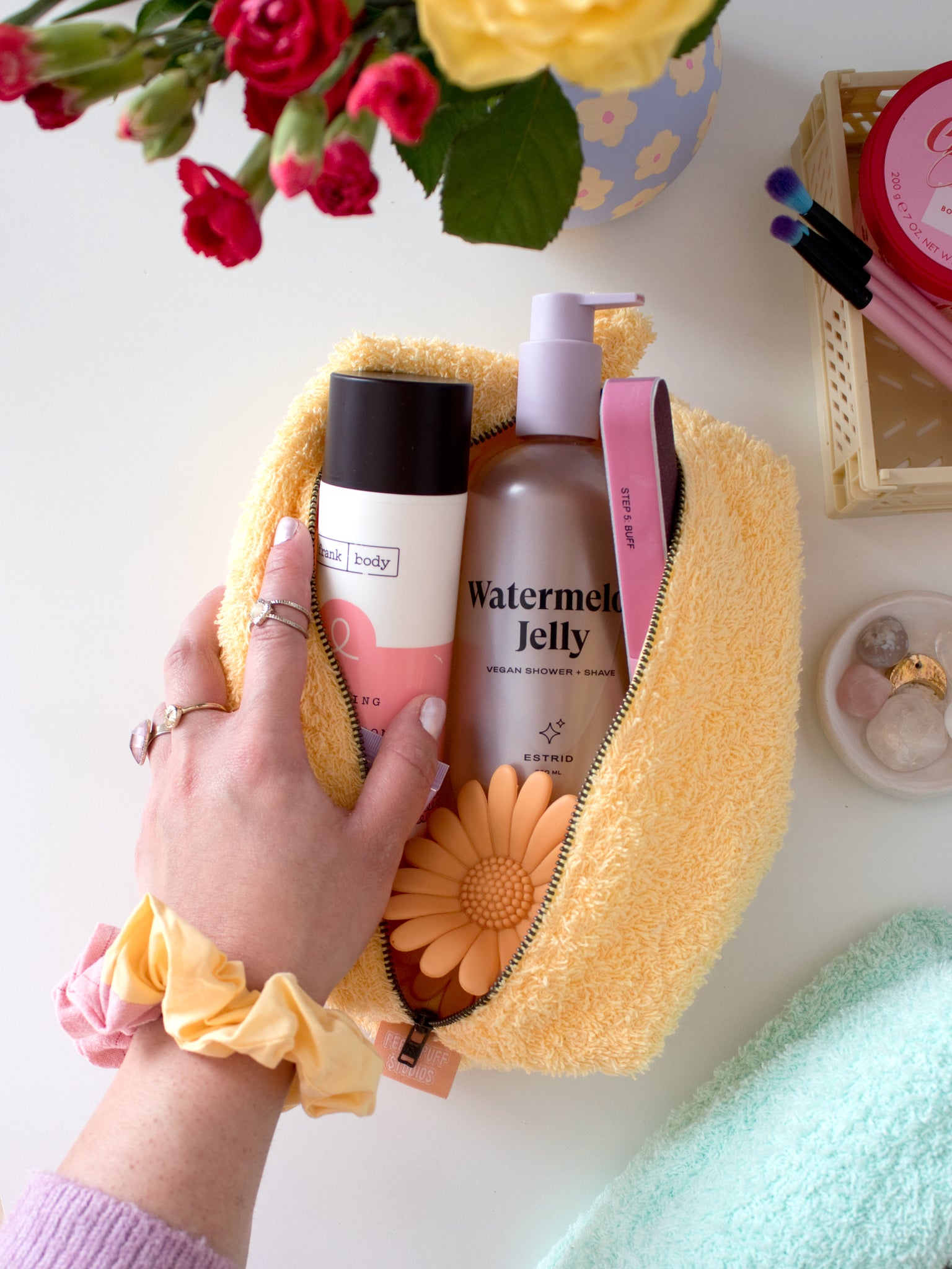 A female hand with a scrunchie on her wrist opens a yellow towelling makeup bag to reveal beauty products.