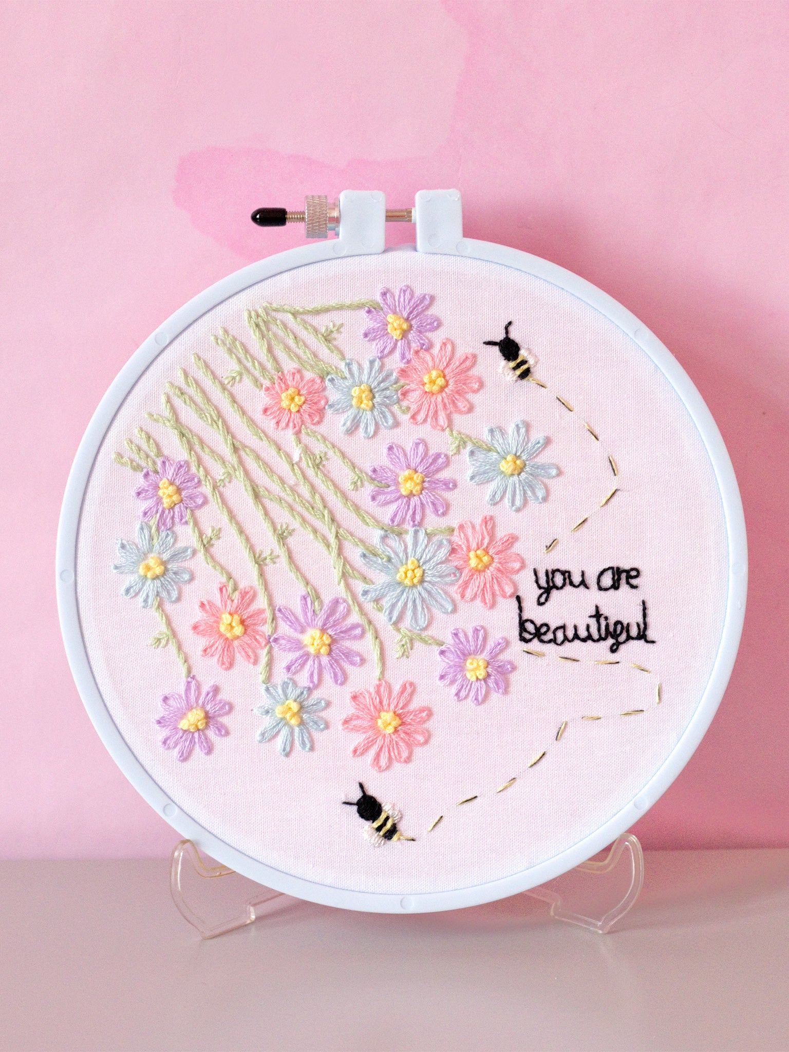 A blue embroidery hoop with white fabric stitched with flowers, bees, and text that reads you are beautiful. Displayed in front of a pink-marbled wall.