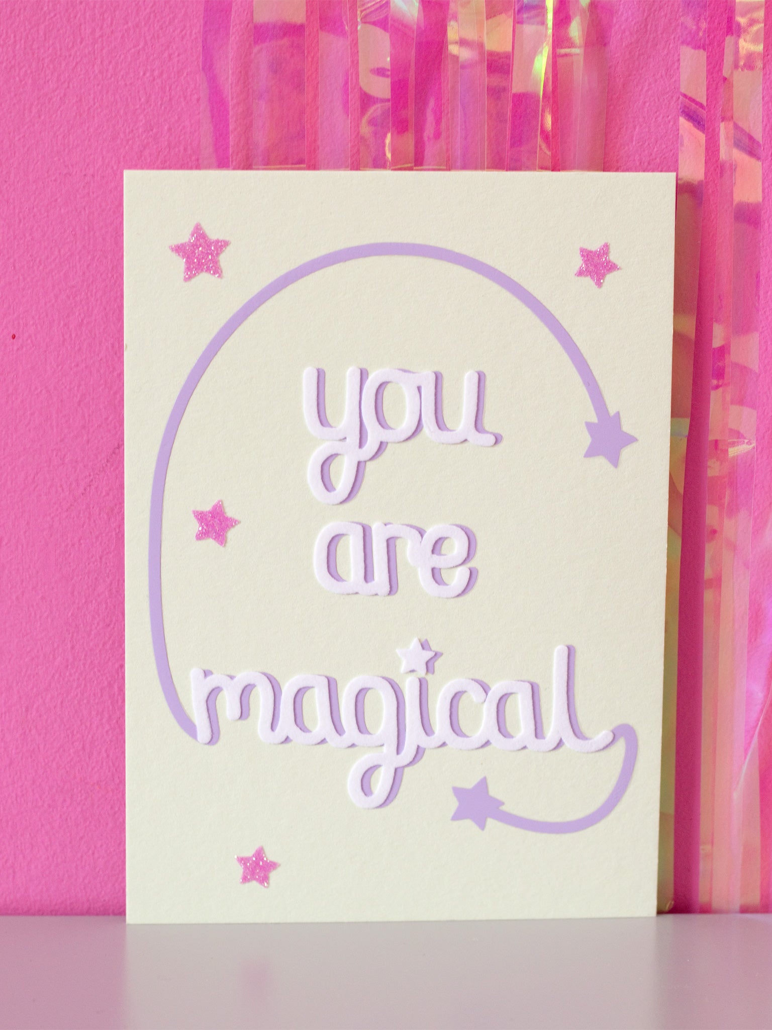 Art print with You Are Magical in flocked font with a purple curved line with a star at the end, coming off with M and L of magical, sitting in front of a pink wall.