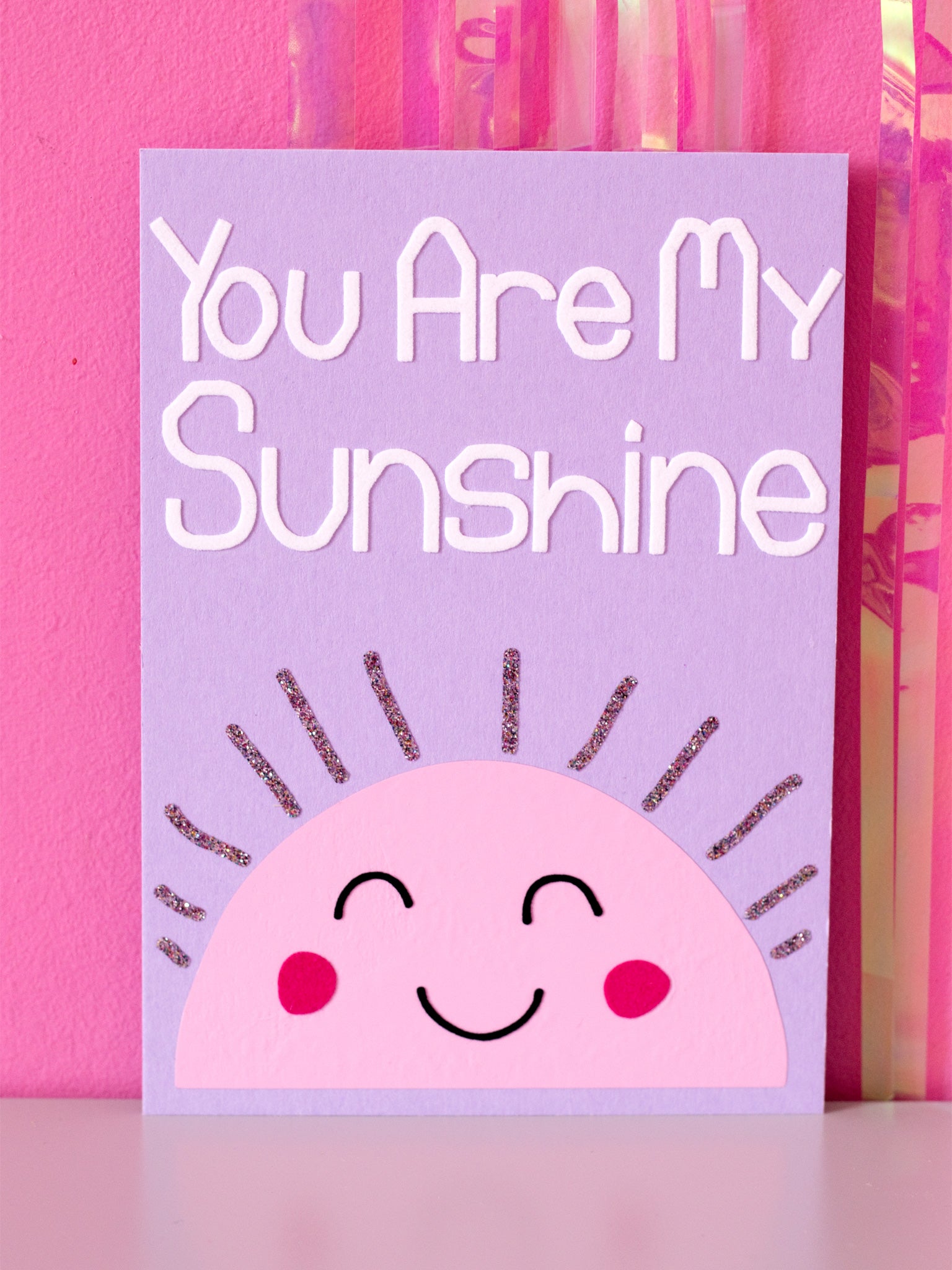 Art print with You Are My Sunshine in flocked font on top of a pink, simple drawn sun, with glittery beams and a cute face, sitting in front of a pink wall.