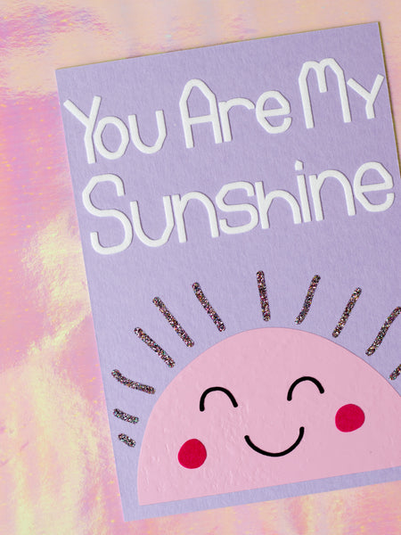 Art print with You Are My Sunshine in flocked font on top of a pink, simple drawn sun, with glittery beams and a cute face.